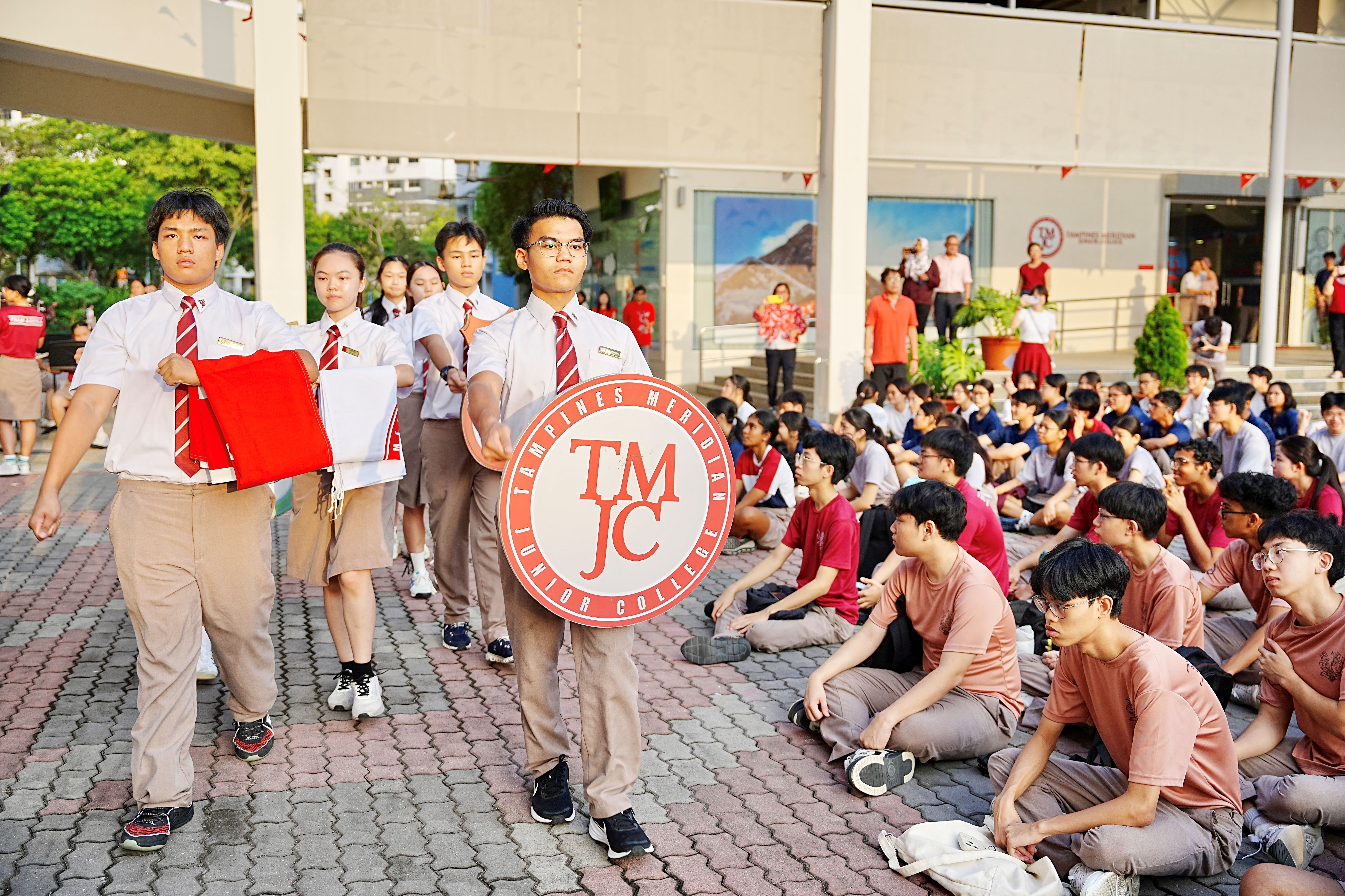 TMJC students in school uniforms march in procession across an outdoor courtyard, with the lead student carrying the TMJC crest, as hundreds of fellow students sit on the ground watching.